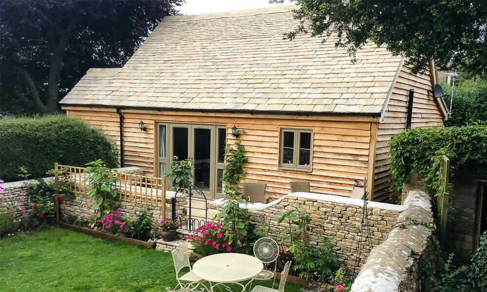 Aldridge Annexe with slate roof and wooden walls, overlooking walled garden with flower beds and lawn, and metal patio set in corner